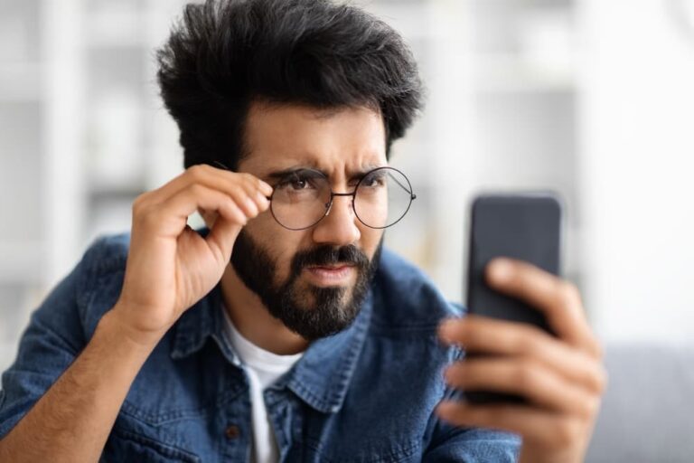 Homem com óculos forçando a visão para enxergar a tela do celular, ilustrando os sintomas de astigmatismo.