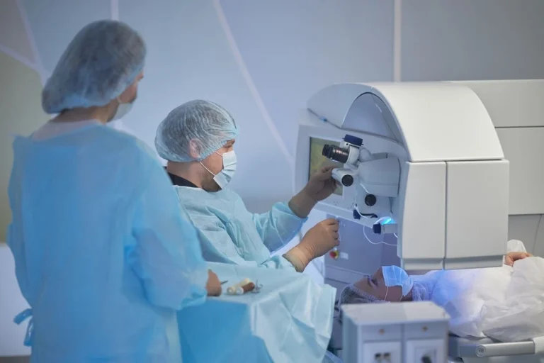 Paciente deitado durante uma cirurgia LASIK para correção da miopia, com um aparelho de laser posicionado sobre o rosto e dois oftalmologistas acompanhando o procedimento.